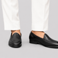 A pair of black leather slip-on loafers worn with white trousers, shown from the side against a solid gray background.
