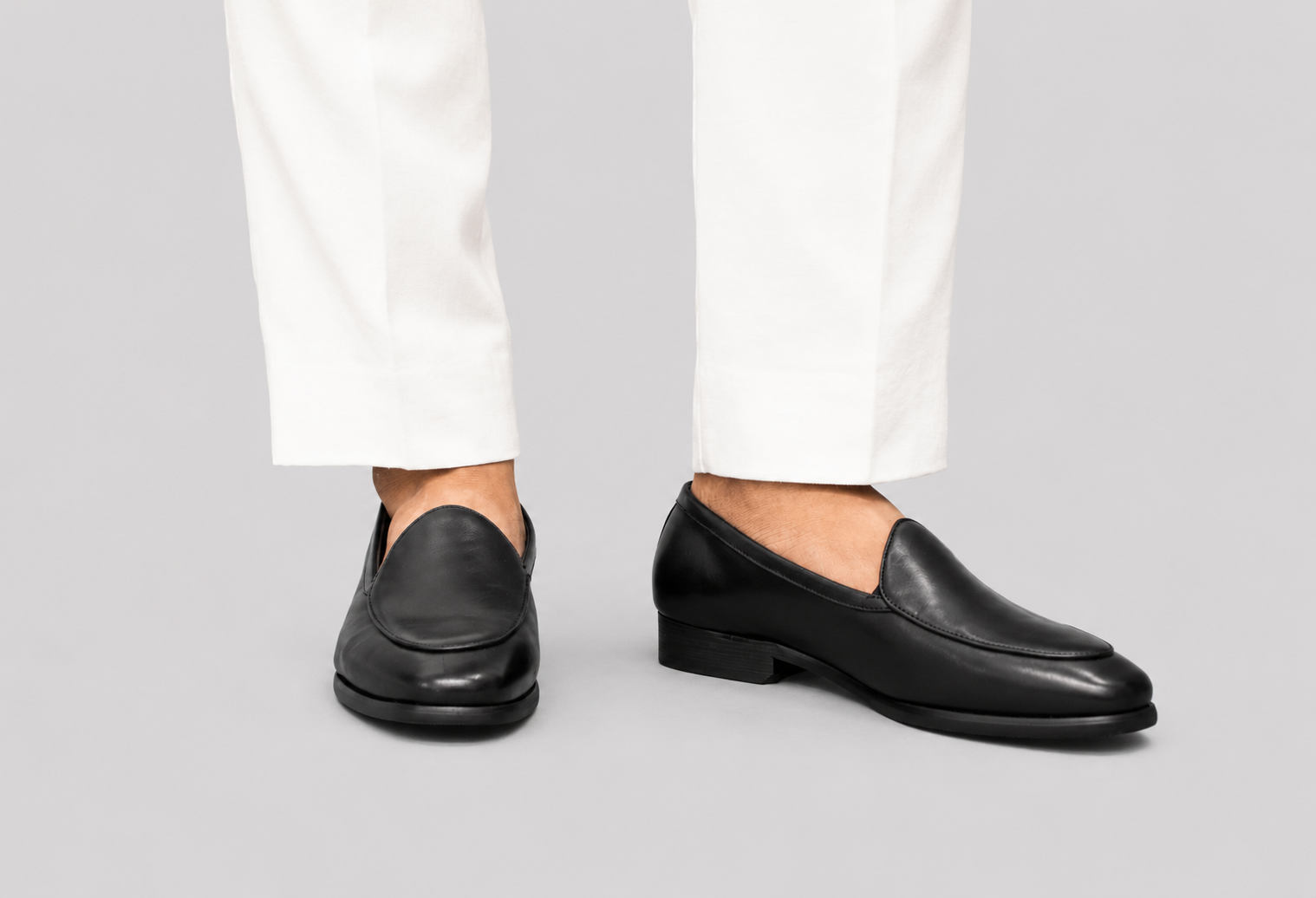 A pair of black leather slip-on loafers worn with white trousers, shown from the side against a solid gray background.