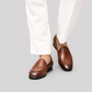 A person wearing brown leather venetian loafers with white trousers, shown from the knees down against a solid light gray background.
