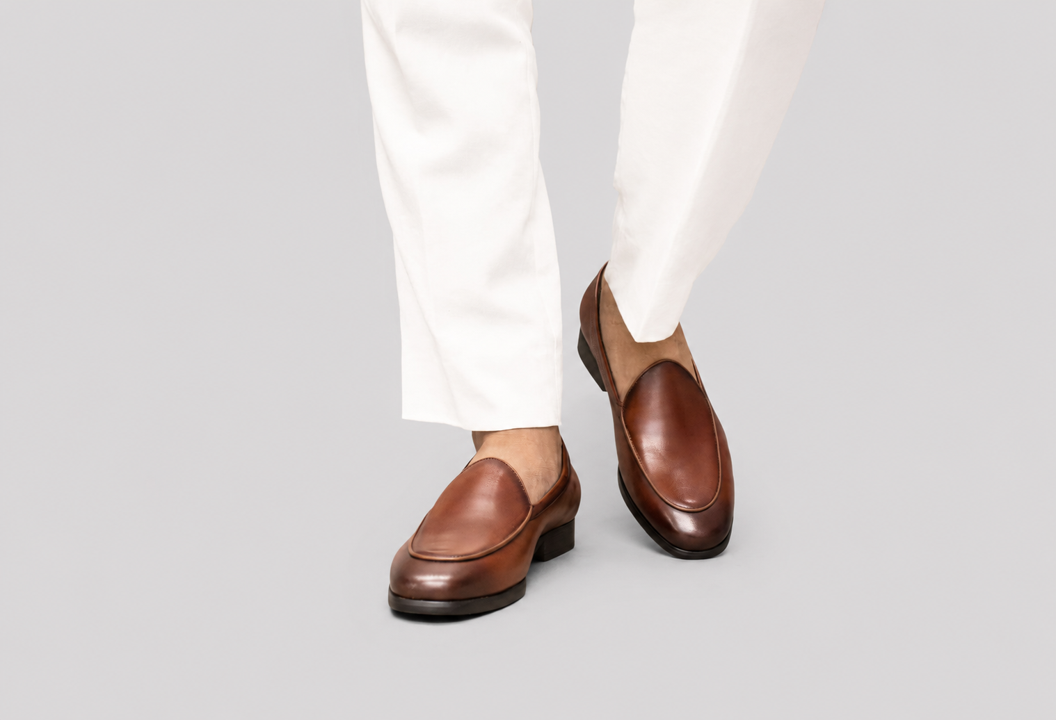 A person wearing brown leather venetian loafers with white trousers, shown from the knees down against a solid light gray background.