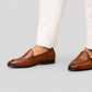 A pair of men's brown leather venetian loafers worn with white trousers, shown from the side against a solid light gray background.