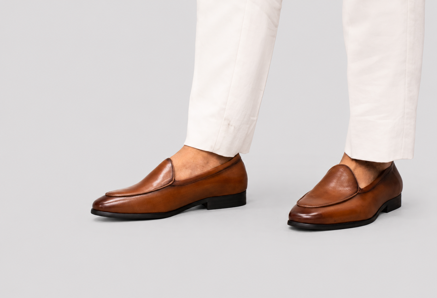 A pair of men's brown leather venetian loafers worn with white trousers, shown from the side against a solid light gray background.