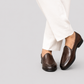 A person wearing dark brown leather loafers with white trousers, shown from the knees down against a solid light gray background.