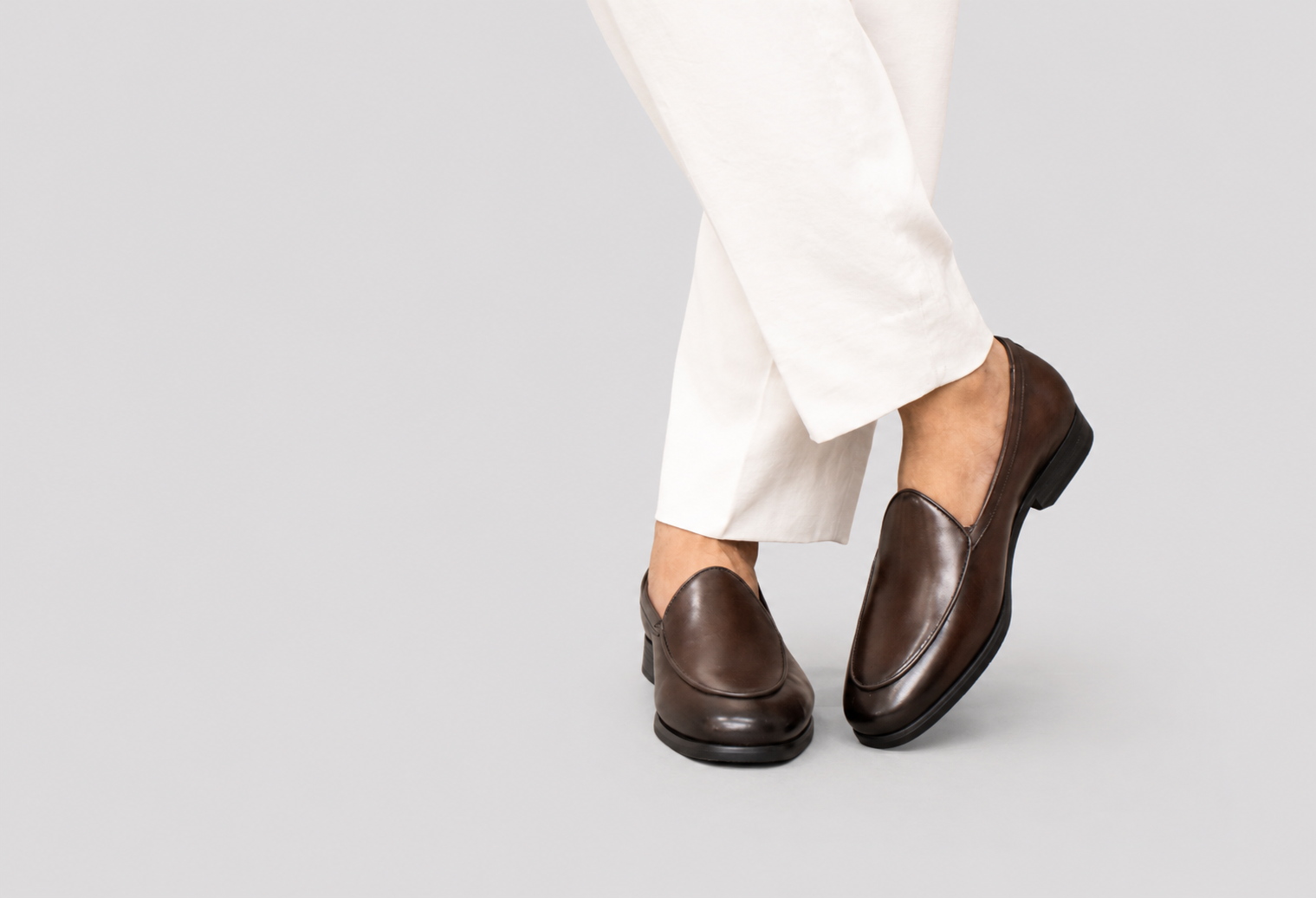 A person wearing dark brown leather loafers with white trousers, shown from the knees down against a solid light gray background.